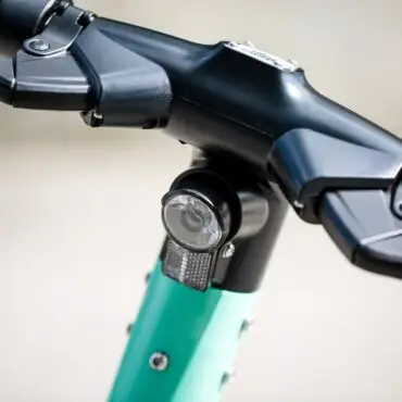 Close-up of a black bicycle handlebar stem with a circular front light mounted on the teal frame area beneath.