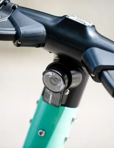 Close-up of a black bicycle handlebar stem with a circular front light mounted on the teal frame area beneath.