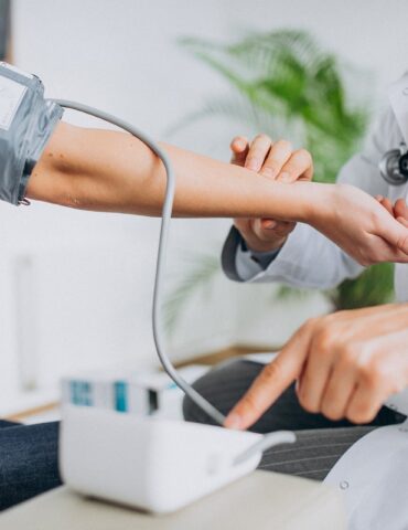 Doctor checks patient’s blood pressure using a cuff; a stethoscope rests around the doctor’s neck.