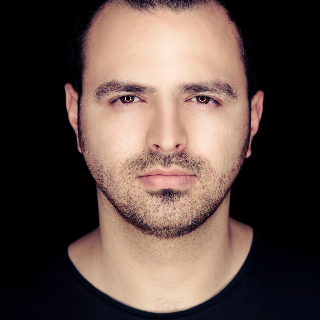 Close-up portrait of a man with short dark hair and light stubble against a black background, looking straight at the camera with a neutral expression.