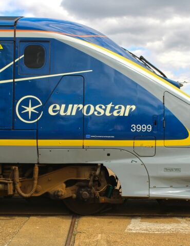Blue Eurostar high-speed train side with white logo and branding, at a station platform with people in the background.