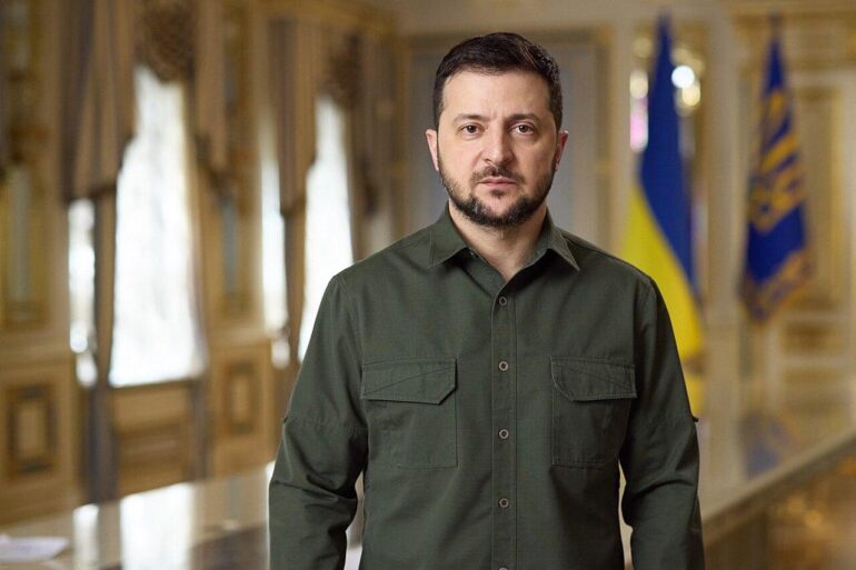 Man in an olive green button-down shirt stands in a grand, gold-toned hallway with flags visible in the background.