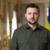 Man in an olive green button-down shirt stands in a grand, gold-toned hallway with flags visible in the background.