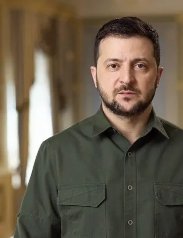 Man in an olive green button-down shirt stands in a grand, gold-toned hallway with flags visible in the background.