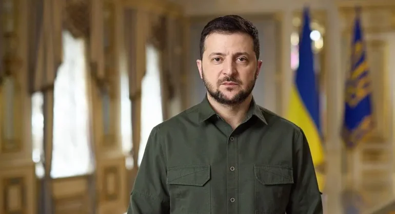 Man in an olive green button-down shirt stands in a grand, gold-toned hallway with flags visible in the background.