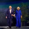 Older man in a dark suit and pink tie walks beside an older woman in a royal blue pantsuit on a stage at night.