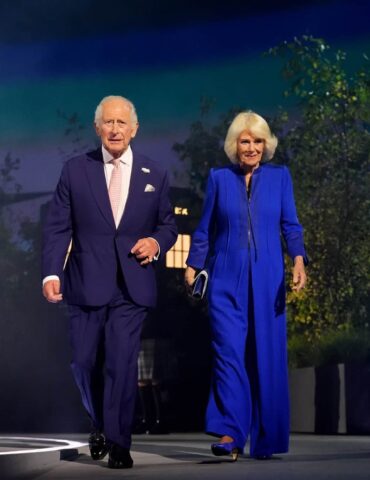 Older man in a dark suit and pink tie walks beside an older woman in a royal blue pantsuit on a stage at night.