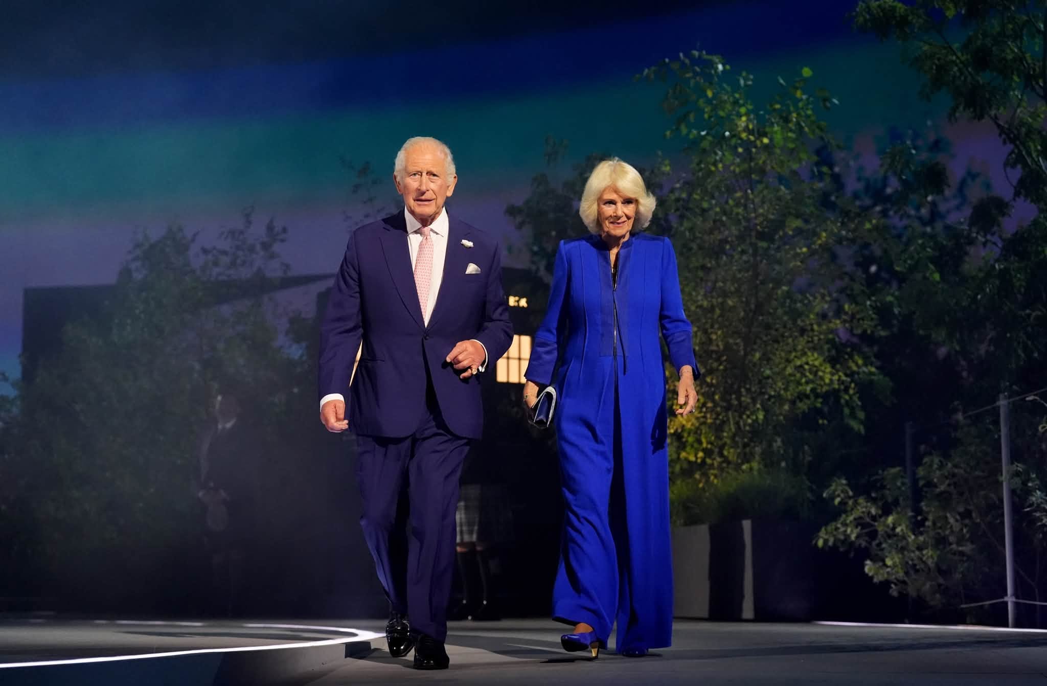 Older man in a dark suit and pink tie walks beside an older woman in a royal blue pantsuit on a stage at night.