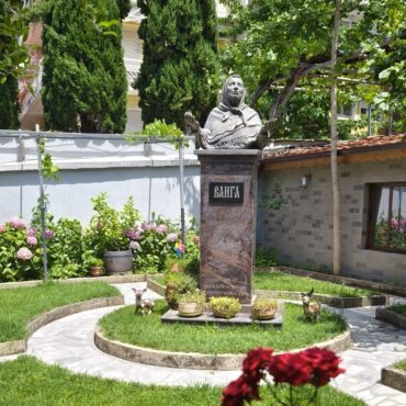 Stone statue of a veiled woman atop a pedestal in a small garden with green shrubs and pink hydrangeas; decorative deer statues nearby.