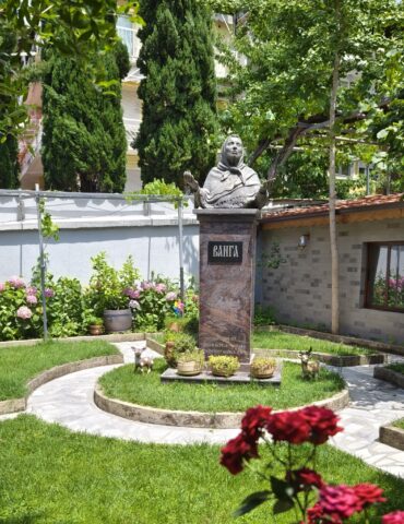 Stone statue of a veiled woman atop a pedestal in a small garden with green shrubs and pink hydrangeas; decorative deer statues nearby.