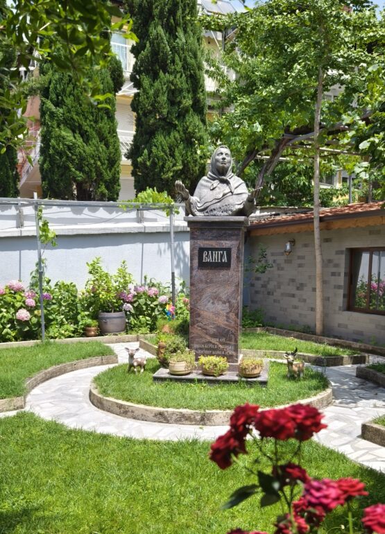 Baba_Vanga_bust - NOTE.radio - Polskie radio w UK | Polskie radio w Londynie Stone statue of a veiled woman atop a pedestal in a small garden with green shrubs and pink hydrangeas; decorative deer statues nearby.