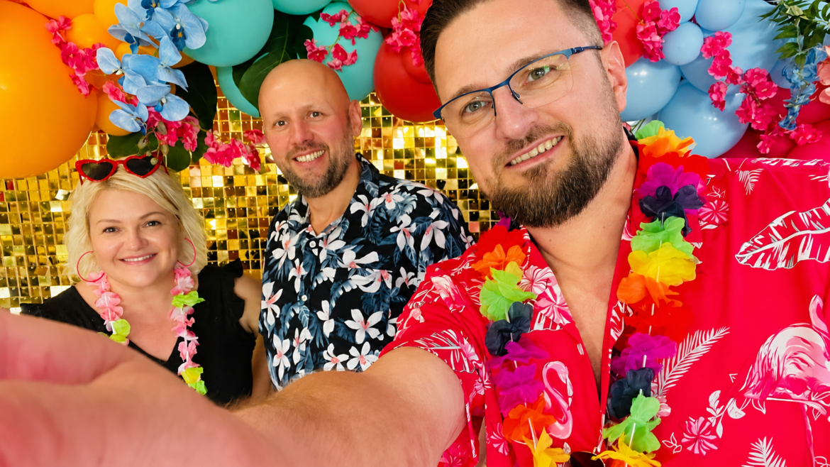 Group selfie at a tropical party with colorful balloons, flowers, and a gold sequin backdrop behind three smiling friends in Hawaiian shirts and leis.