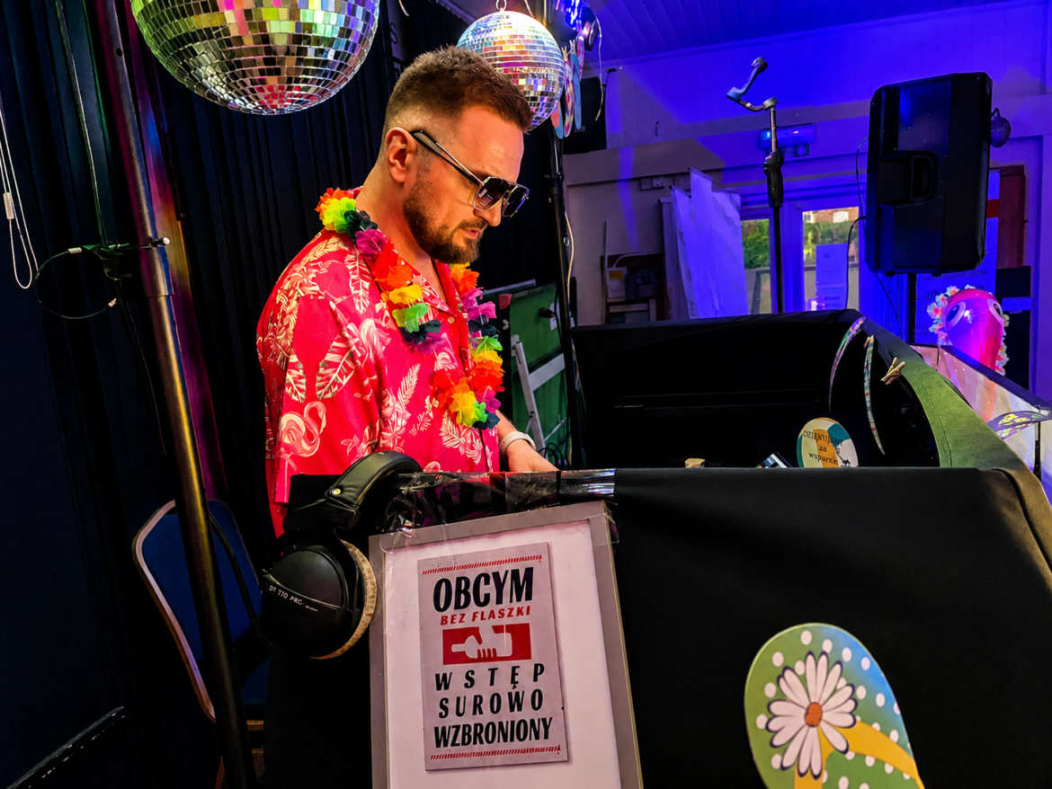 DJ in a pink tropical shirt and flower leis performing at a colorful party, wearing sunglasses and headphones at the turntables.