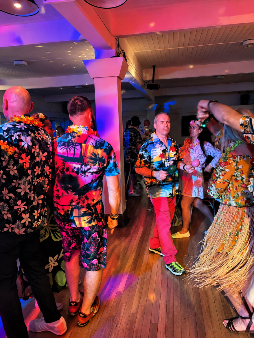 Group of people in bright tropical shirts dancing and mingling at a Hawaiian-themed party under colorful lighting.