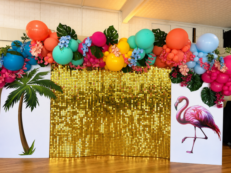 Aloha party - NOTE.radio - Polskie radio w UK | Polskie radio w Londynie Tropical party backdrop with a bright balloon arch over a gold sequin wall, plus palm leaf cutouts and flamingo artwork on the sides.