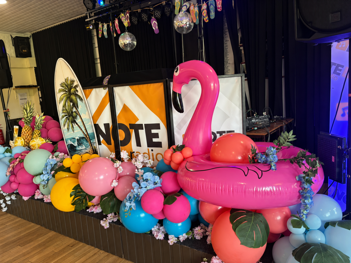 Stage decorated for a tropical party with a pink flamingo float, palm leaves, and colorful balloons beside a Note Radio banner.