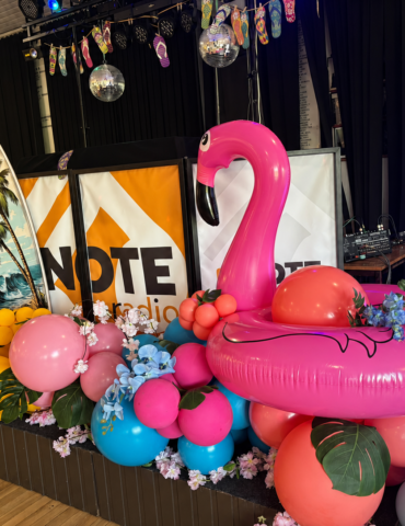 Stage decorated for a tropical party with a pink flamingo float, palm leaves, and colorful balloons beside a Note Radio banner.