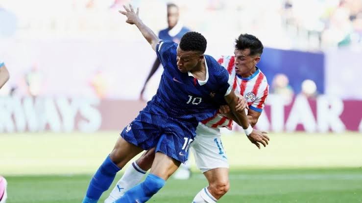 Francja Paragwaj - NOTE.radio - Polskie radio w UK | Polskie radio w Londynie Two soccer players battle for the ball during a match, blue jersey shielding the ball from a defender in red-and-white stripes.