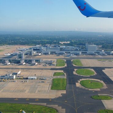 Aerial view of a busy airport: terminal buildings, runways, and several parked airplanes with a blue airplane wing in the top right corner.