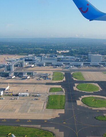 Aerial view of a busy airport: terminal buildings, runways, and several parked airplanes with a blue airplane wing in the top right corner.