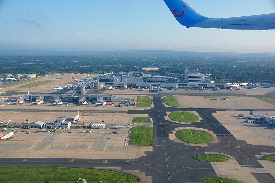 Aerial view of a busy airport: terminal buildings, runways, and several parked airplanes with a blue airplane wing in the top right corner.