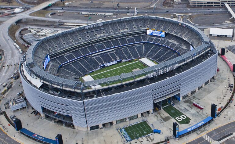 Aerial view of MetLife Stadium with a football field inside the bowl and surrounding seating and structures outside the arena.