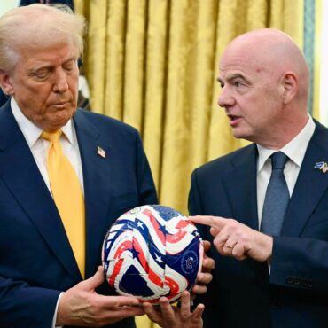 Two men in dark suits hold a red, white, and blue star-spangled ball indoors, one touching it as they discuss it.