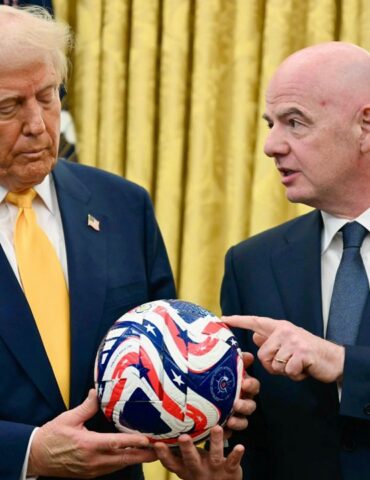 Two men in dark suits hold a red, white, and blue star-spangled ball indoors, one touching it as they discuss it.