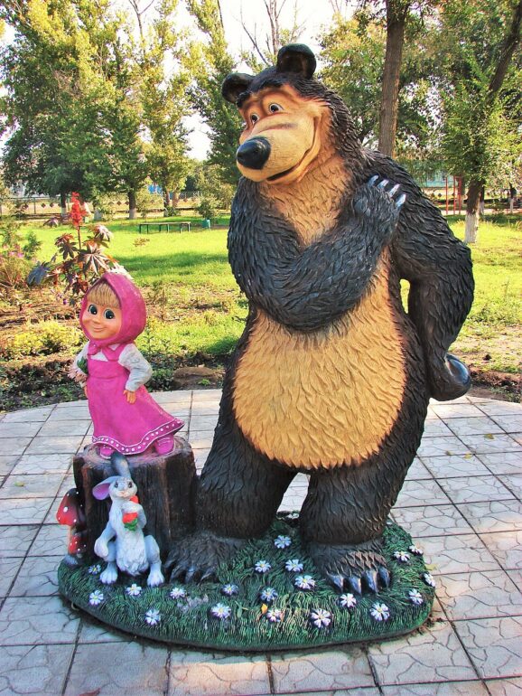 Large bear statue in a park with a small girl statue in a pink hood and a rabbit nearby on a flowered pedestal.