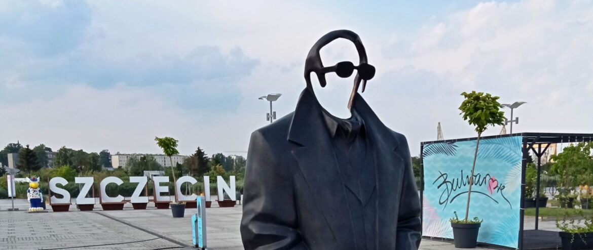 Statue of a person in a black suit with round head and sunglasses, standing outdoors with a briefcase; large SZCZECIN letters in the background.