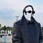 Statue of a person in a black suit with round head and sunglasses, standing outdoors with a briefcase; large SZCZECIN letters in the background.