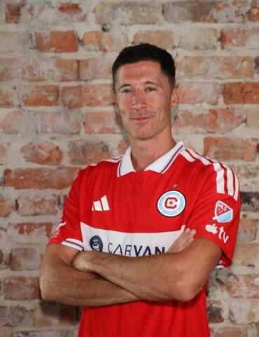 Portrait of a man in a red soccer jersey with arms crossed against a brick wall background.