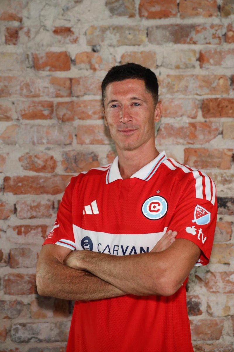 Portrait of a man in a red soccer jersey with arms crossed against a brick wall background.