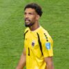 Soccer goalkeeper in a yellow jersey with number 1, walking on a green pitch, focused expression and team crest on the chest.