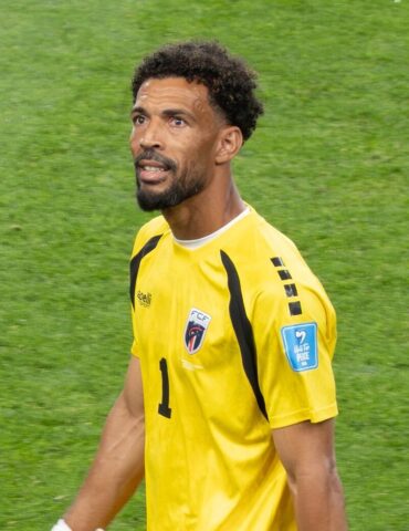 Soccer goalkeeper in a yellow jersey with number 1, walking on a green pitch, focused expression and team crest on the chest.