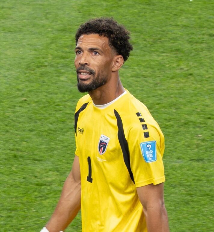 Soccer goalkeeper in a yellow jersey with number 1, walking on a green pitch, focused expression and team crest on the chest.