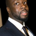 Close-up portrait of a Black man in a navy suit and white shirt, with a full beard, looking slightly to the side against a dark background.