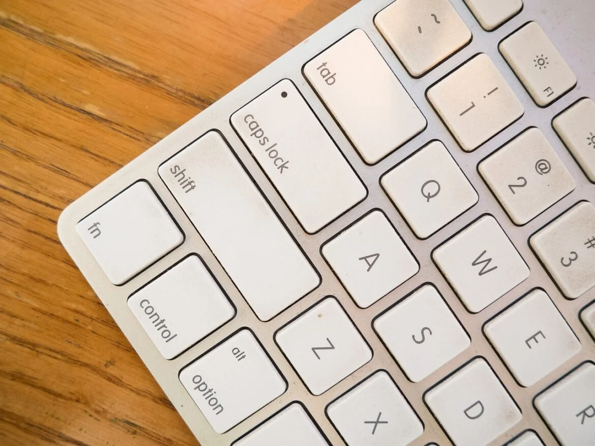 Close-up of a white keyboard on a wooden desk, showing keys such as Caps Lock, Tab, Shift, Control, and letter keys A, W, S, Z.