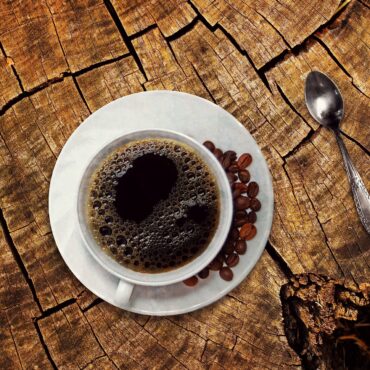 Top-down view of a coffee cup on a white saucer with coffee beans, resting on a cracked wooden tree stump; a silver spoon lies to the right.