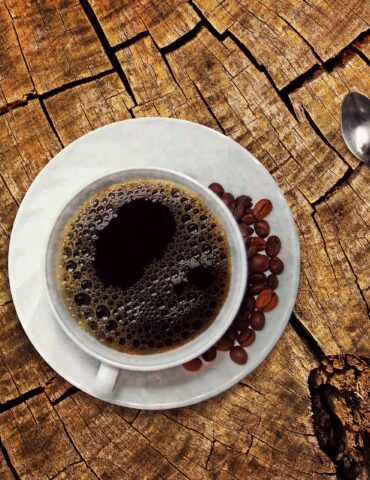 Top-down view of a coffee cup on a white saucer with coffee beans, resting on a cracked wooden tree stump; a silver spoon lies to the right.