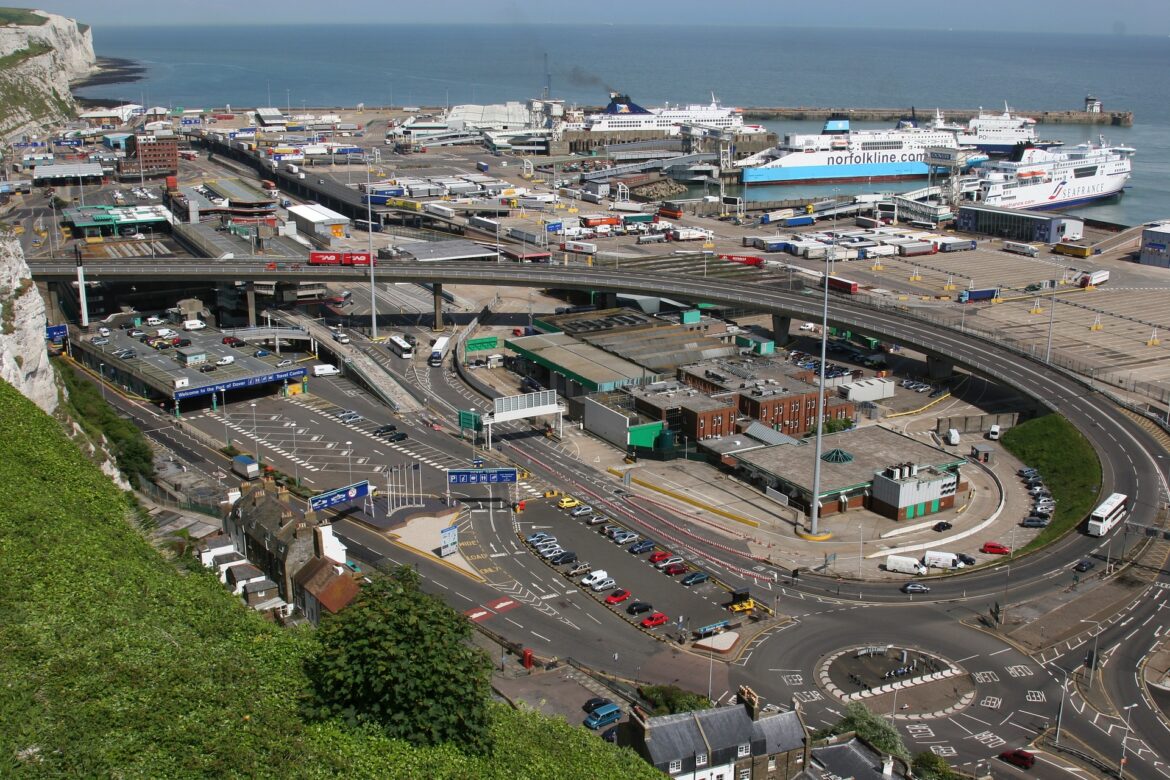 A busy coastal port with ferries, trucks, and roads, set against the sea and white cliffs in the background.