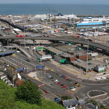 A busy coastal port with ferries, trucks, and roads, set against the sea and white cliffs in the background.