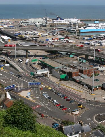 A busy coastal port with ferries, trucks, and roads, set against the sea and white cliffs in the background.