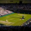 Tennis match on a sunlit grass court with a player serving, surrounded by a packed stadium.