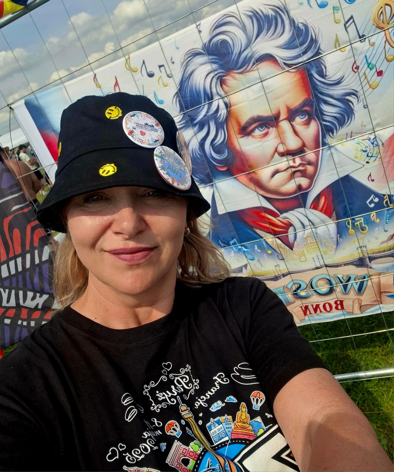 Close-up selfie of a smiling woman wearing a black bucket hat with two pins, in front of a vibrant mural featuring a portrait of Albert Einstein and musical notes.