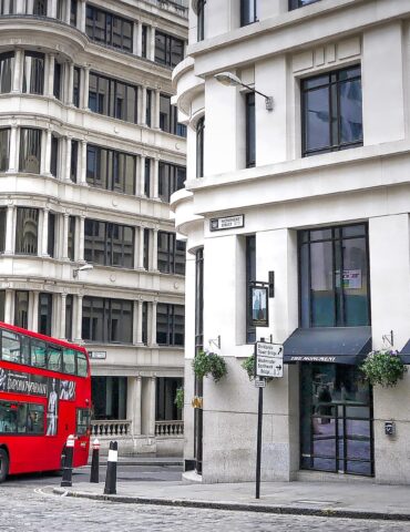 Red double-decker bus on a cobblestone street beside white curved-edge buildings with tall windows in a cityscape