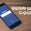 Phone shows Facebook login screen beside Scrabble tiles spelling SOCIAL MEDIA on a wooden surface.
