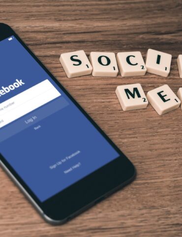 Phone shows Facebook login screen beside Scrabble tiles spelling SOCIAL MEDIA on a wooden surface.