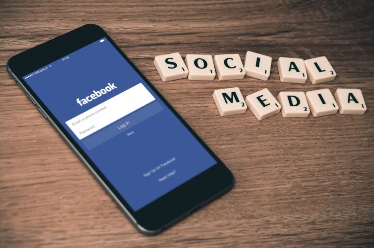 Phone shows Facebook login screen beside Scrabble tiles spelling SOCIAL MEDIA on a wooden surface.