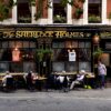 Exterior of The Sherlock Holmes pub with golden signage, black facade, and outdoor dining along a brick street. Plants hang from above.